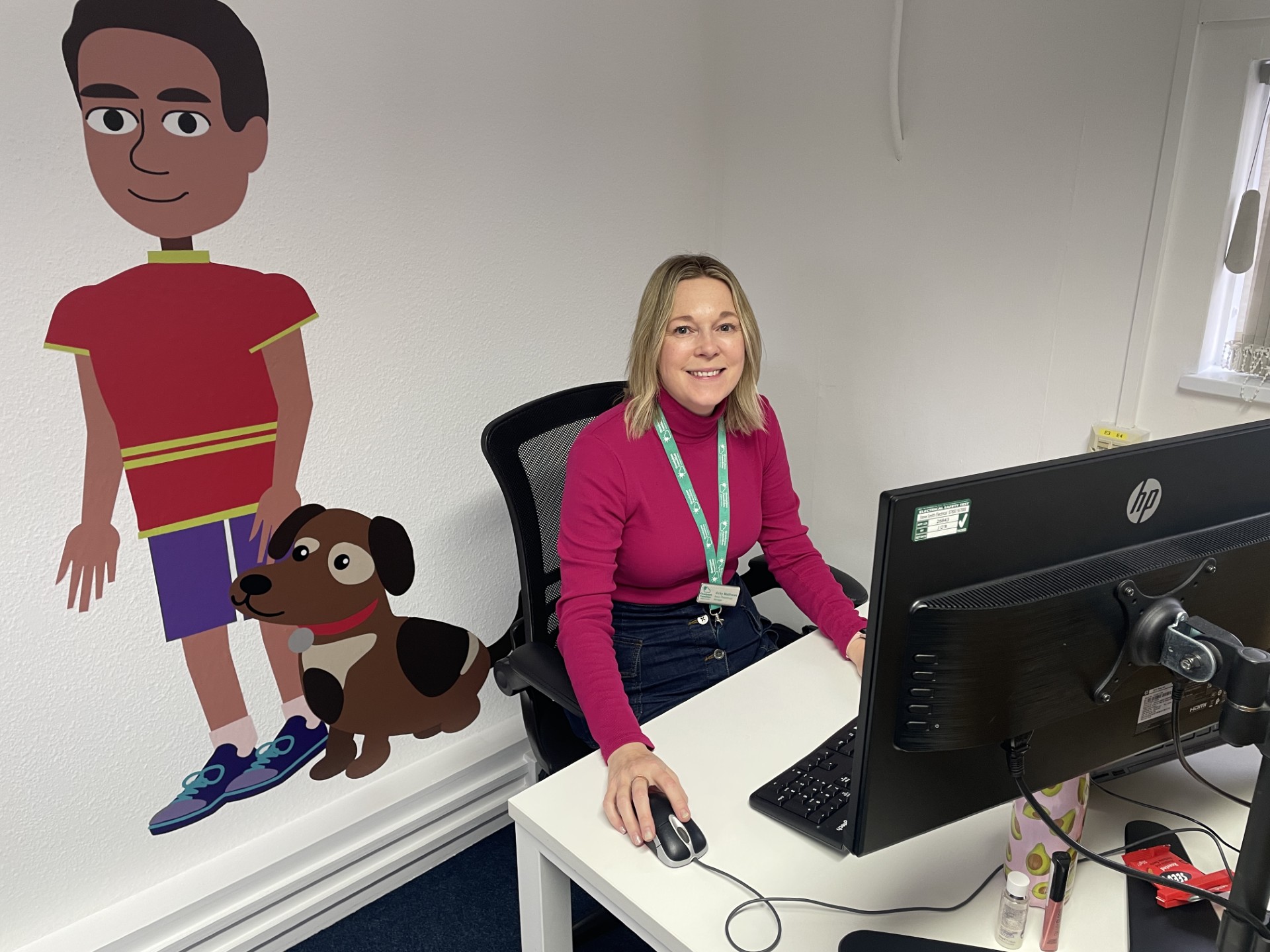 A woman sits at a desk and smiles. An animated logo is printed on the wall beside her.